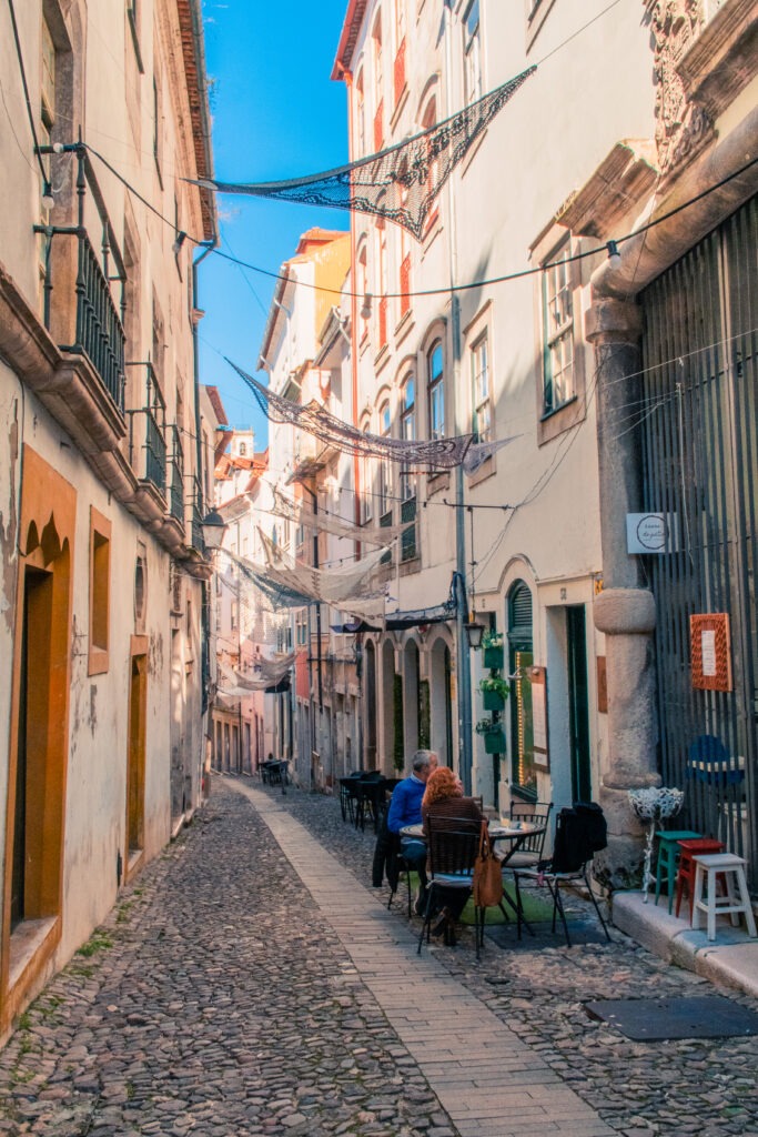 cobblestone street lined with buildings with sails overhead and couple eating at table and chairs in Coimbra