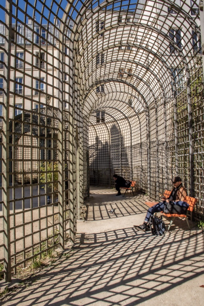 Arched metal trellis walkway in Jardin Anne Frank casts geometric shadows across the path while visitors relax on orange benches in the sunlight. This peaceful corner of Jardin Anne Frank offers a quiet retreat in the heart of Paris surrounded by greenery and historic architecture.