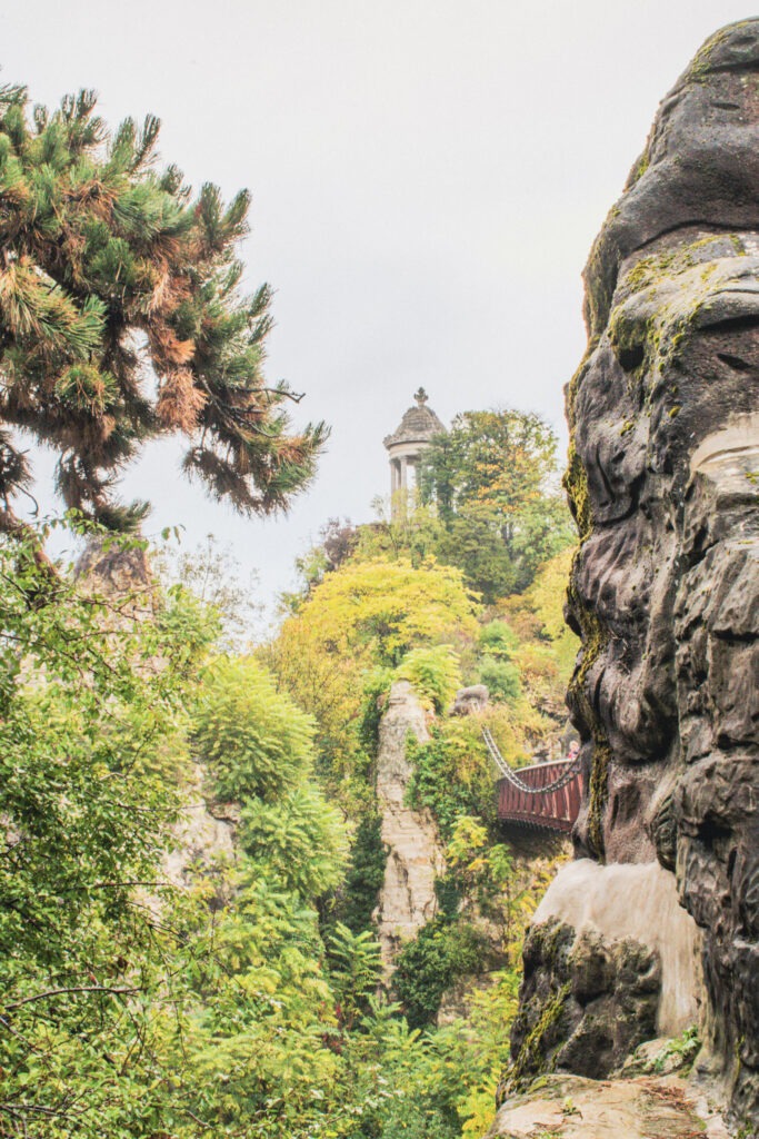 Alt text: Lush greenery, dramatic rock formations, and a suspended footbridge lead through a scenic landscape in Parc des Buttes Chaumont, with a hilltop temple visible in the distance. This unique park is one of the most memorable gardens to visit in Paris, offering winding paths, panoramic views, and a peaceful escape from the city's busy streets.