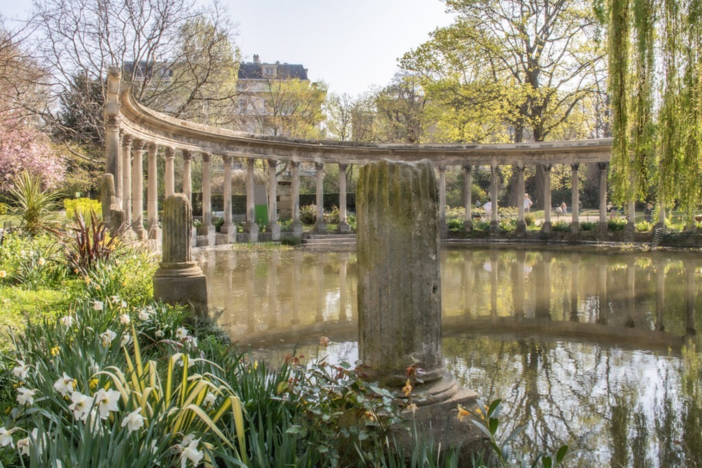 Curved stone colonnade reflected in a peaceful pond surrounded by spring flowers, lush greenery, and mature trees in a historic Paris garden. This tranquil scene showcases why Paris gardens to visit are among the city's most relaxing attractions, offering quiet paths, elegant landscaping, and hidden architectural details.