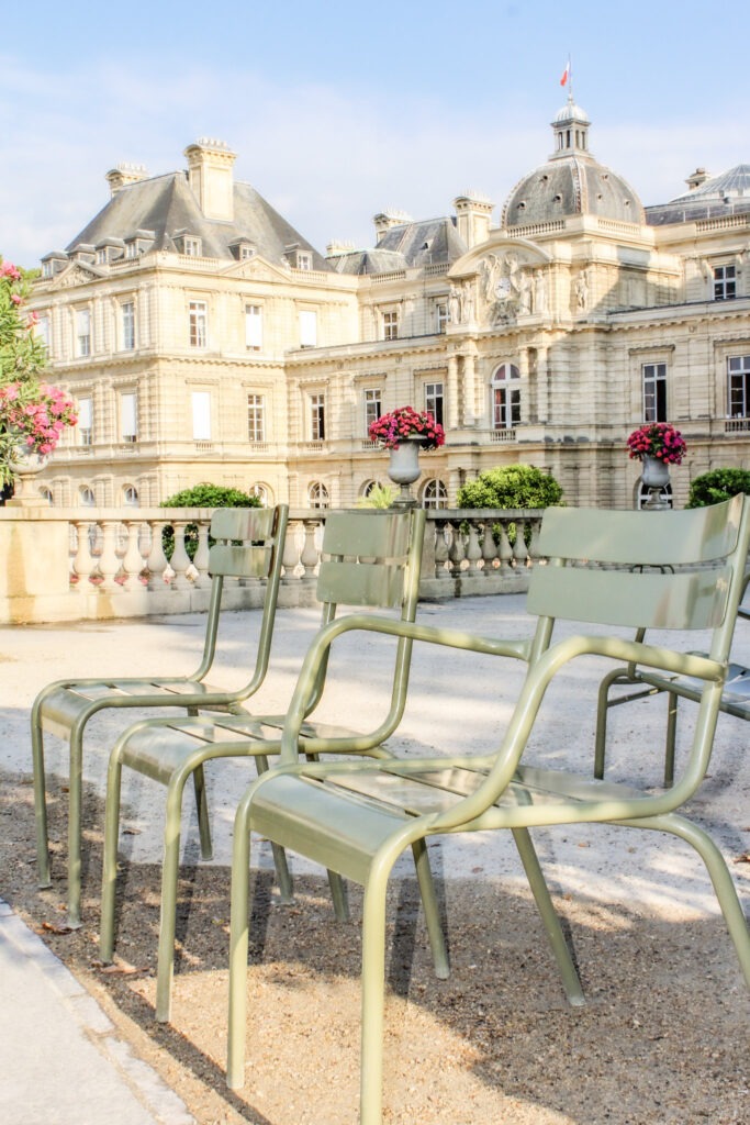 Green chairs line a gravel pathway beside colorful flower beds and historic buildings in Luxembourg Gardens. Relaxing in one of the best gardens in Paris