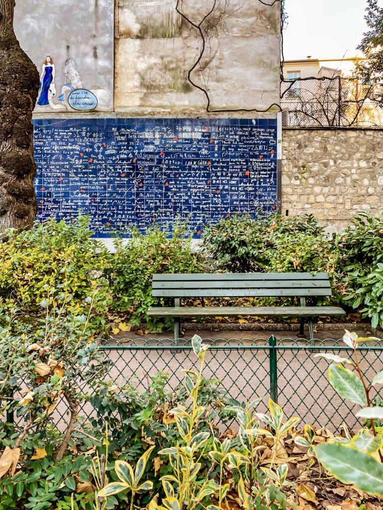 Green park bench surrounded by shrubs faces the Wall of Love in Paris, a blue tiled mural covered with handwritten messages in dozens of languages. The quiet garden setting makes this one of the most romantic places in Paris for couples seeking a peaceful spot away from the crowds.