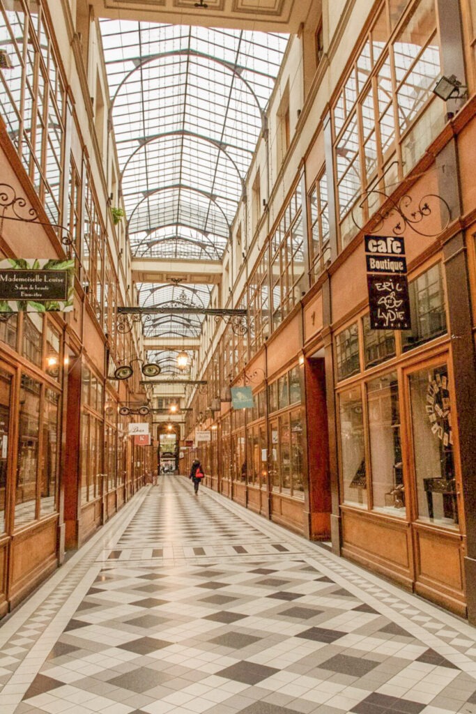 Elegant covered passage in Paris with a glass vaulted ceiling, patterned tile floor, and boutique storefronts lining both sides. The soft light and intimate atmosphere make this historic arcade a romantic place in Paris for a leisurely stroll together.