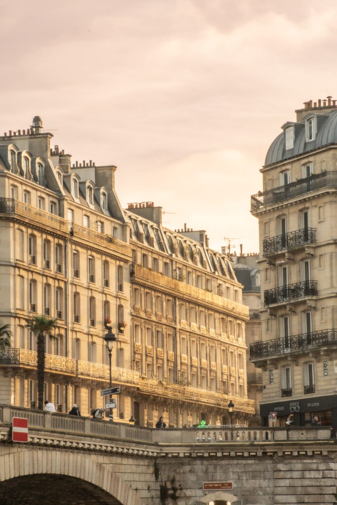 Sunlight glows across elegant Paris apartment buildings lining a river canal at sunset. Pedestrians walk along the stone bridge below, capturing the romantic architecture and atmosphere that make Paris worth visiting.