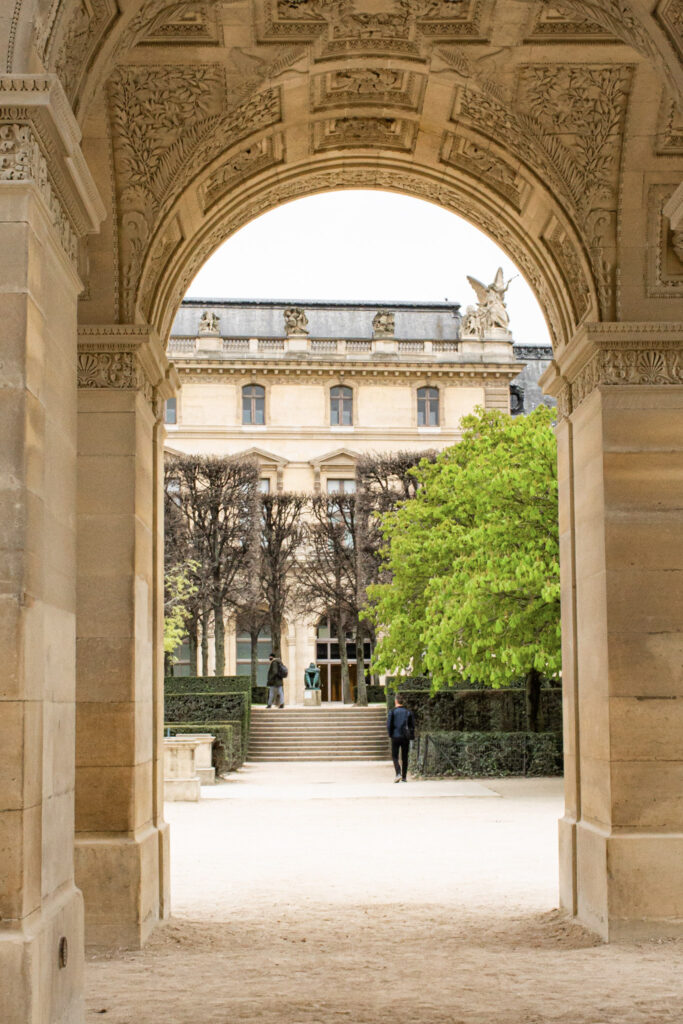 7 Hour Layover in Paris: Can You Leave the Airport? 2 Ornate stone archway opens into a quiet courtyard garden surrounded by historic Paris architecture and trimmed trees. A few people walk through the peaceful space, highlighting one of the reasons why people like to do a 7 hours layover in Paris
