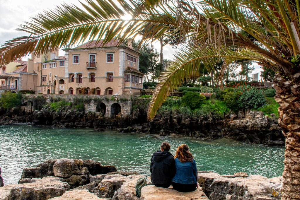 couple sitting on rock under palm tree by sea on day trips lisbon