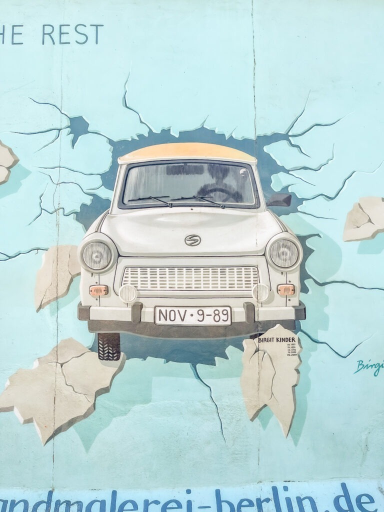wall mural in berlin showing car coming through berlin wall when comparing berlin to paris