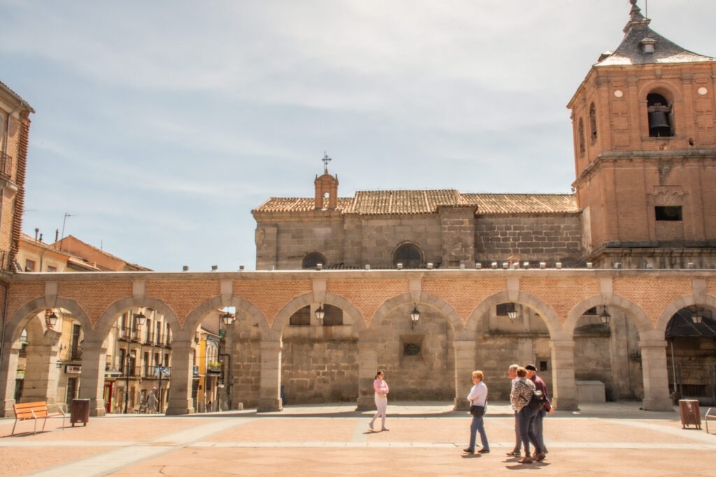 large square with people walking with several stone archways and tall clock tower on a avila day trip from madrid