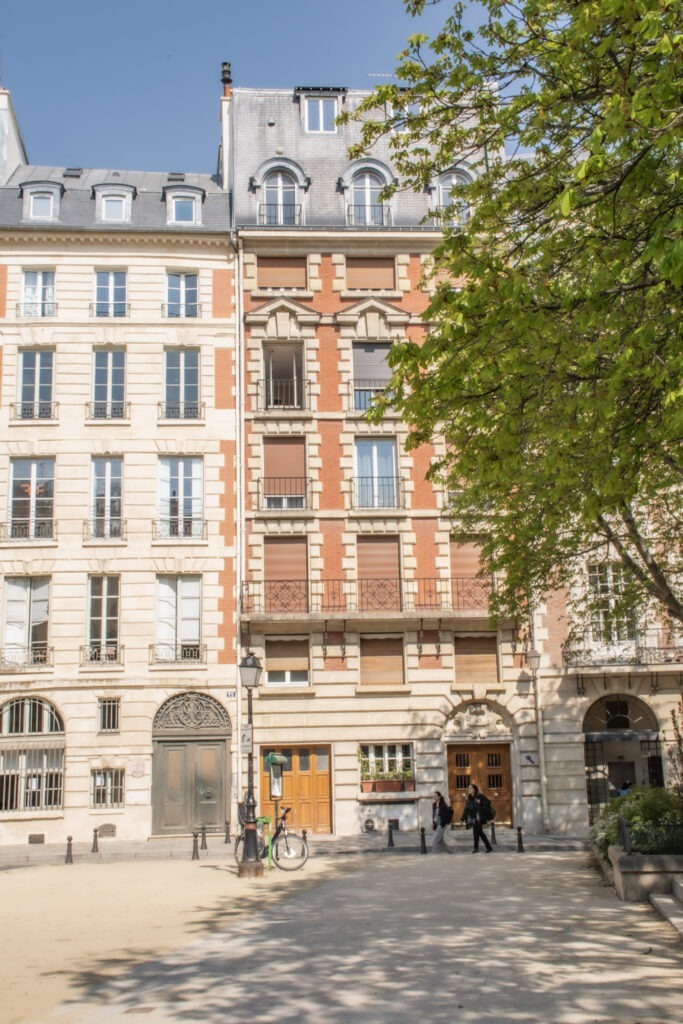 Classic Paris apartment buildings with cream stone and red brick facades overlook a quiet tree lined square. A bicycle rests near the walkway as a few pedestrians pass by, reflecting the everyday neighborhood moments that make slow travel in Paris so rewarding.
