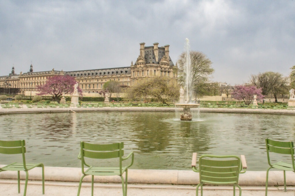 Alt text: Green chairs surround a large fountain in the Tuileries Garden with the Louvre Palace visible in the background and pink flowering trees framing the scene. Visitors can sit beside the water and linger in the gardens, capturing the relaxed pace of slow travel in Paris.