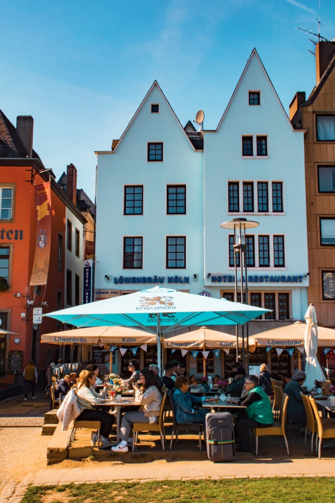 What to See + Do in Cologne in One Day 5 while spending one day in Cologne dine along the Rhine River where under umbrellas there is long tables and chairs with people enjoying food and beer