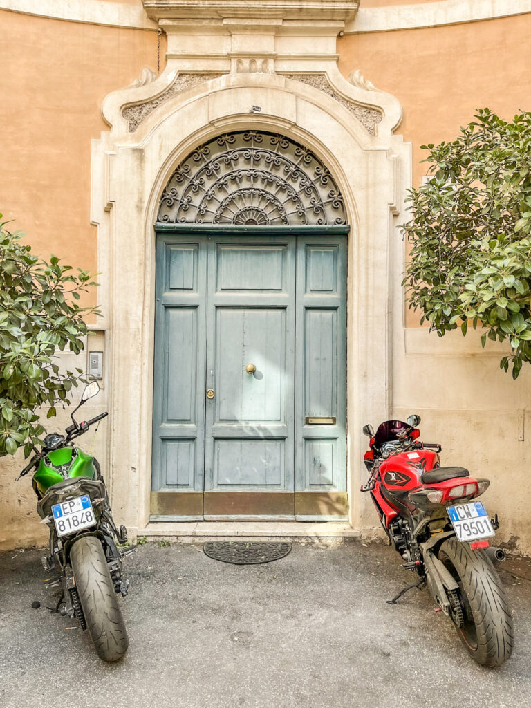a blue door with two motorcycles parked outside with olive trees flanked on either side in rome