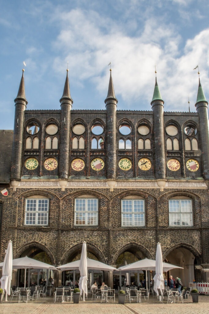 black facade with arches with tables and chairs and umbrellas. the brick building in lubeck has steep towers and regional insignias  