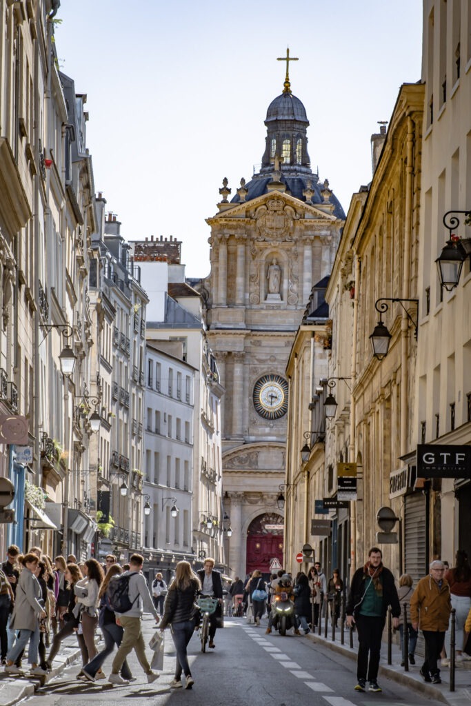 Pedestrians walk along a lively Paris street lined with historic buildings, leading the eye toward a domed church with an ornate clock tower in the distance. Exploring walkable neighborhoods like this offers a glimpse of everyday Paris life and fits naturally into a 4 day itinerary in Paris.