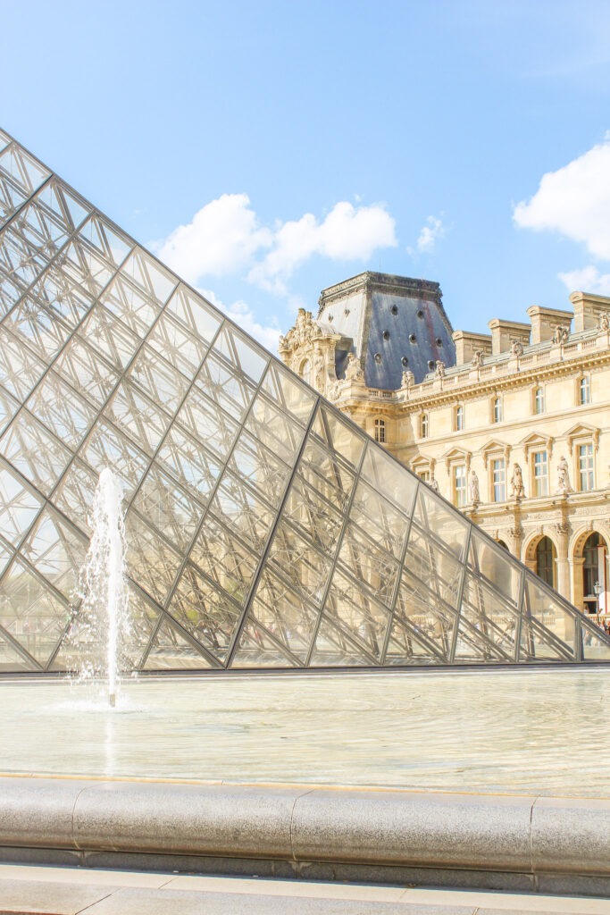 Glass Louvre Pyramid stands beside a fountain with the historic Louvre Palace visible behind it on a sunny day. Exploring the Louvre is a popular highlight for visitors spending 4 days in Paris.
