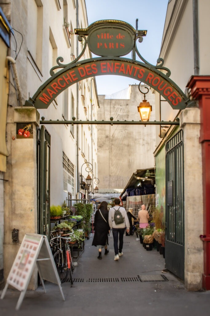 Entrance sign reading “ville de PARIS” and “MARCHÉ DES ENFANTS ROUGES” welcomes visitors into a lively covered market lined with vendors and shoppers. Exploring local markets like this adds a neighborhood experience to a 4 day itinerary in Paris beyond the major attractions.