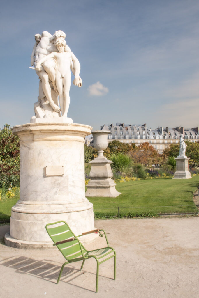 Classical marble statue overlooks manicured gardens and elegant Paris buildings with a green chair resting nearby. Spending time in Paris parks and gardens offers a relaxing break during a 4 day Paris itinerary.