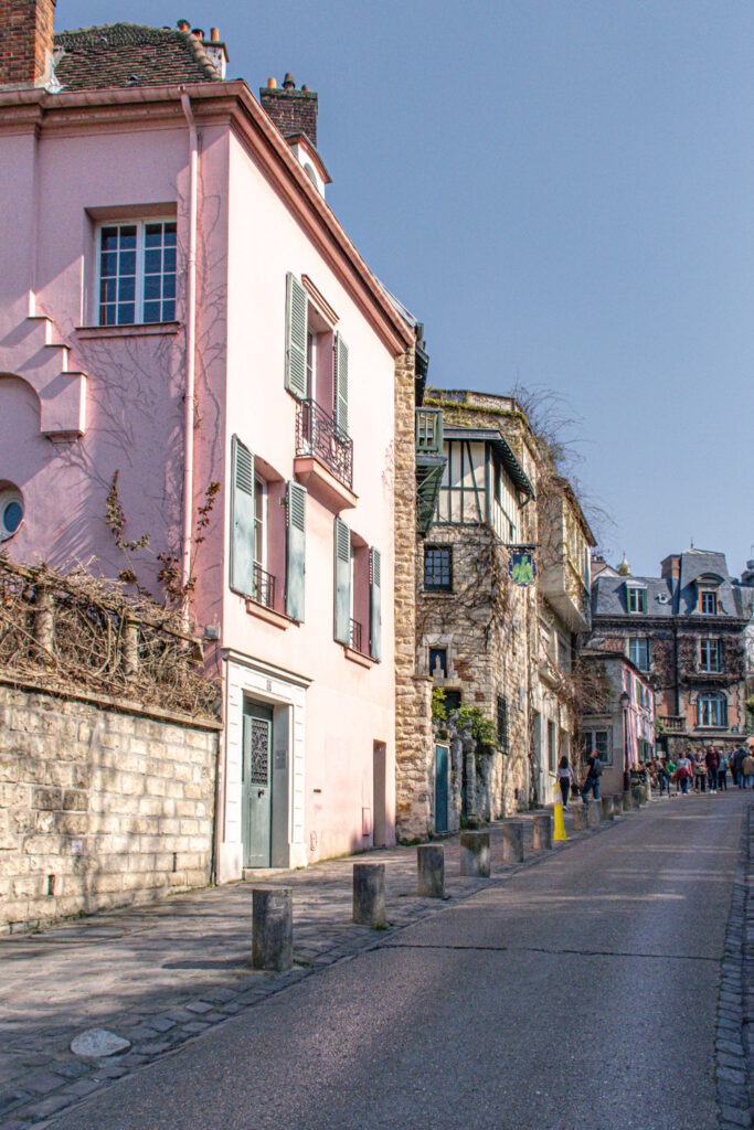 Quiet Montmartre street lined with pastel buildings, stone walls, and historic architecture under a blue sky. Wandering charming neighborhoods like this is one of the highlights of spending 4 days in Paris.