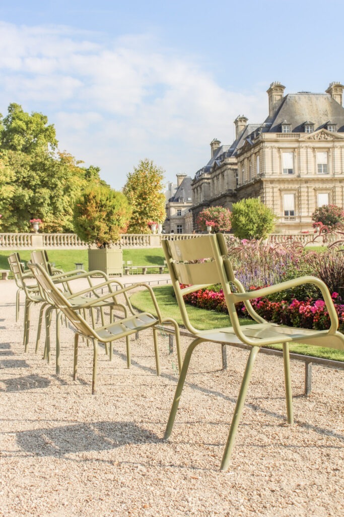 Green chairs line a gravel pathway beside colorful flower beds and historic buildings in Luxembourg Gardens. Relaxing in Paris's famous gardens is an easy way to balance sightseeing during a 4 day Paris itinerary.