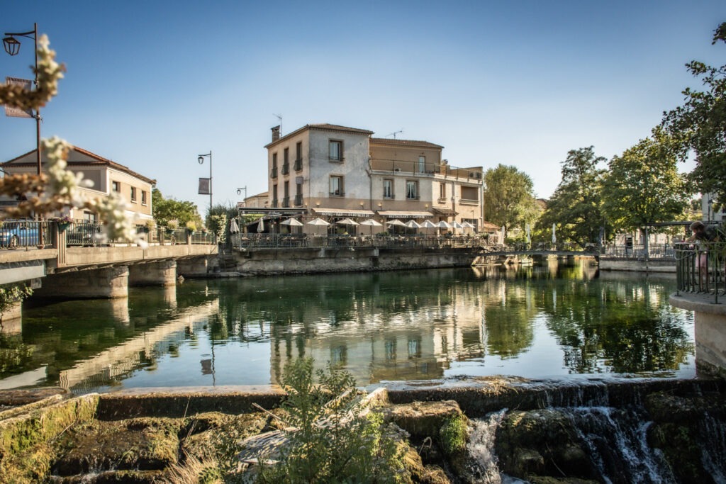 The Sorgue River reflecting waterfront cafés and historic buildings in L’Isle-sur-la-Sorgue, Provence, with a stone bridge and small waterfall in the foreground. The riverside setting is one of the town's main attractions.