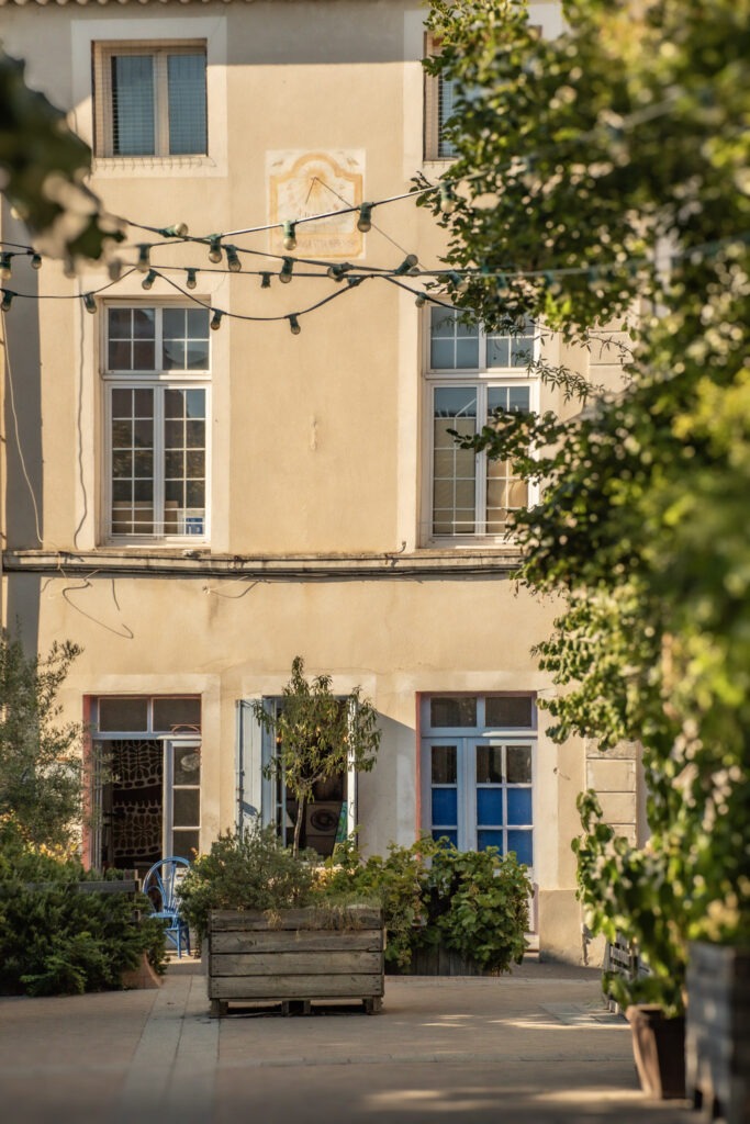 Cream-coloured historic building with shuttered windows, courtyard planters and overhead string lights in L’Isle-sur-la-Sorgue.