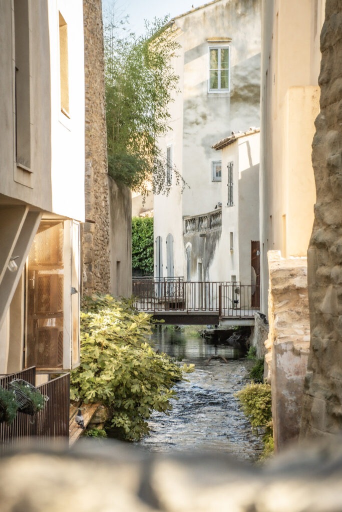 A narrow canal flowing between old stone and stucco buildings, with a small metal footbridge crossing the water. One of the lesser-known waterways running through the historic centre of L’Isle-sur-la-Sorgue, nicknamed the Venice of Provence.
