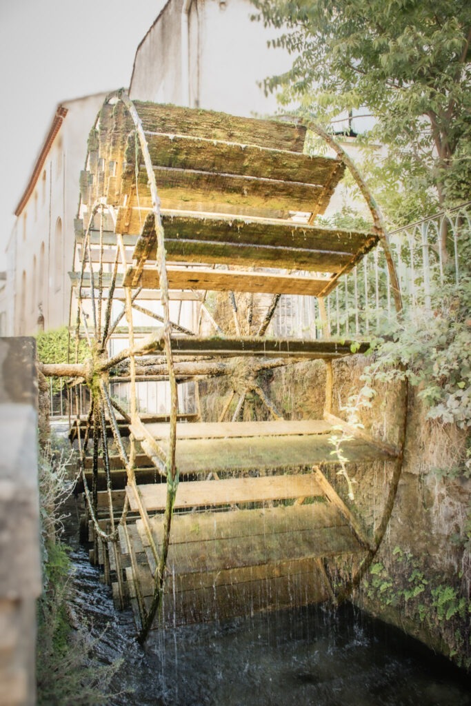 An old wooden waterwheel covered in green moss, with water flowing over its paddles beside a historic building in L’Isle-sur-la-Sorgue. These surviving waterwheels are reminders of the town's former milling and textile industries.