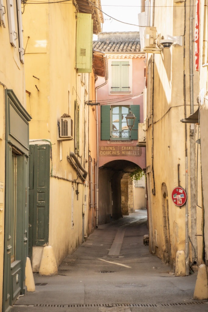 Narrow street in L’Isle-sur-la-Sorgue with pastel buildings and an old archway displaying the faded French words Bains, Chambres, Douches, Meublées.