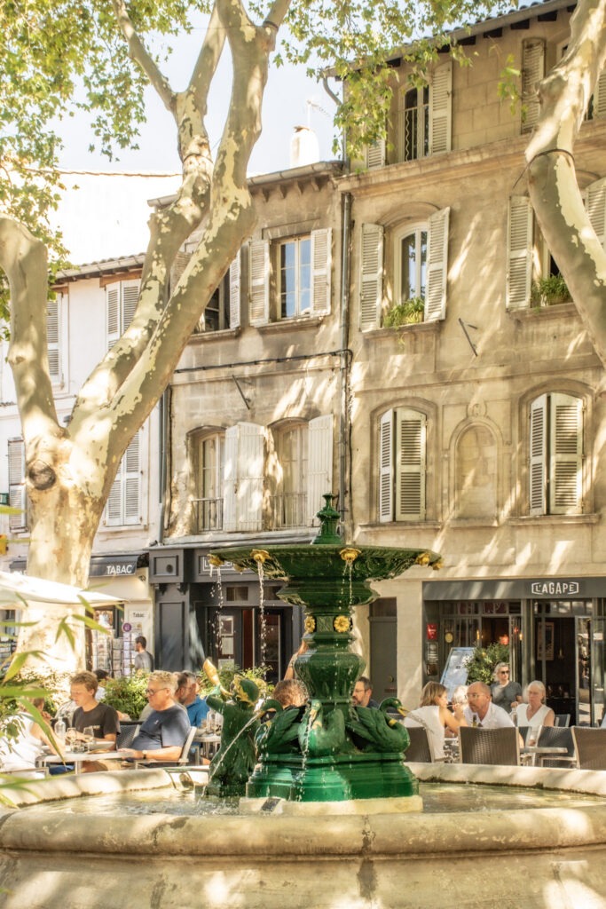 Green fountain surrounded by outdoor restaurant terraces on Place des Corps Saints in Avignon, France.