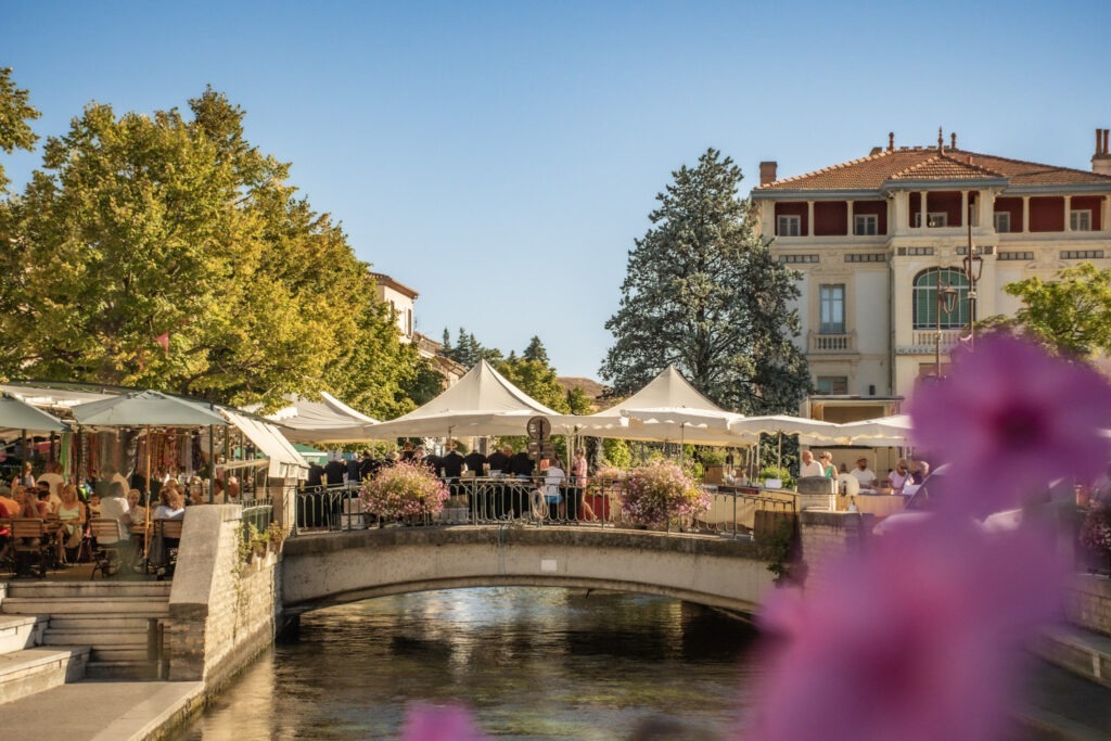Stone footbridge over the Sorgue River with market stalls, people and outdoor café seating in L’Isle-sur-la-Sorgue, Provence.