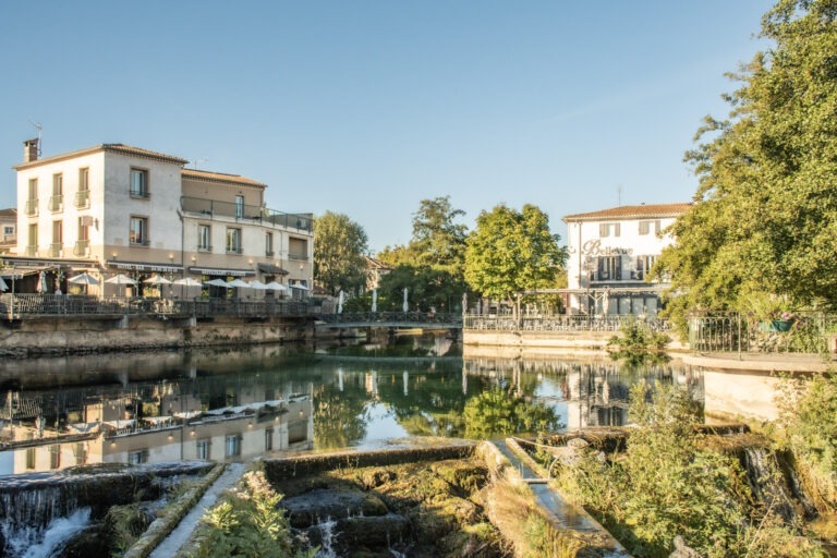Waterfront restaurants and historic buildings reflected in the Sorgue River in L’Isle-sur-la-Sorgue, Provence, on a sunny morning.