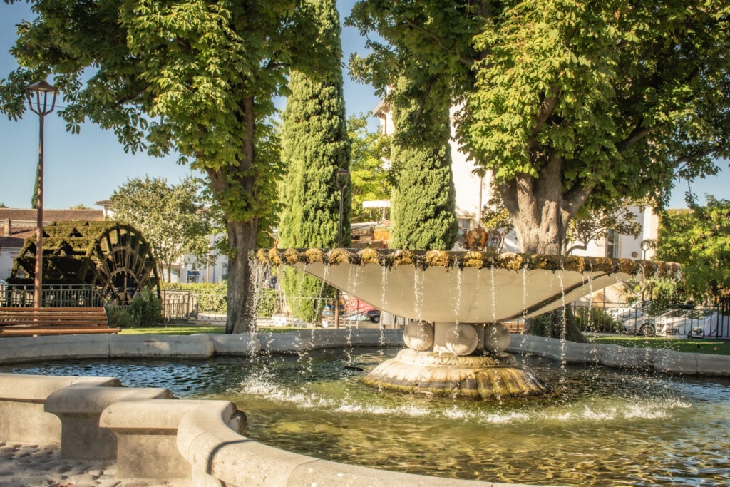 Stone fountain surrounded by trees with a historic waterwheel in the background in L’Isle-sur-la-Sorgue, Provence.