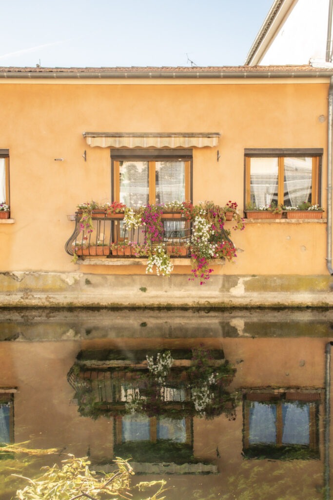 orange stucco building with window and flowers next to canal in L'Isle-sur-la-Sorgue showing water reflections