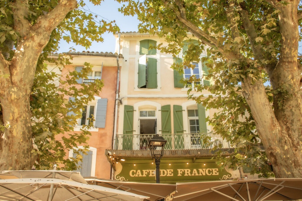 Café de France overlooking Place de la Liberté in L’Isle-sur-la-Sorgue, with green shutters, leafy plane trees and outdoor terrace umbrellas.