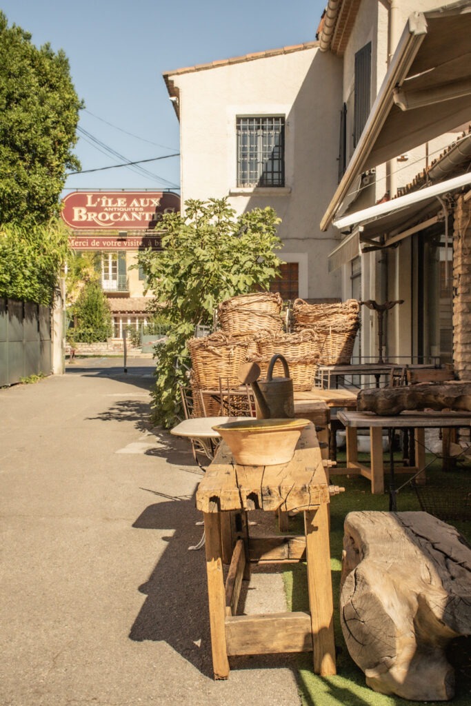 Entrance to L’Île aux Brocantes antique village in L’Isle-sur-la-Sorgue, with vintage wooden furniture and wicker baskets displayed outside.
