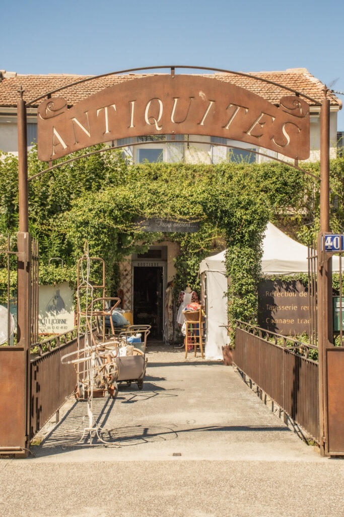 Entrance to an antique dealer courtyard in L’Isle-sur-la-Sorgue, with an iron Antiques sign, vintage furniture and shops.
