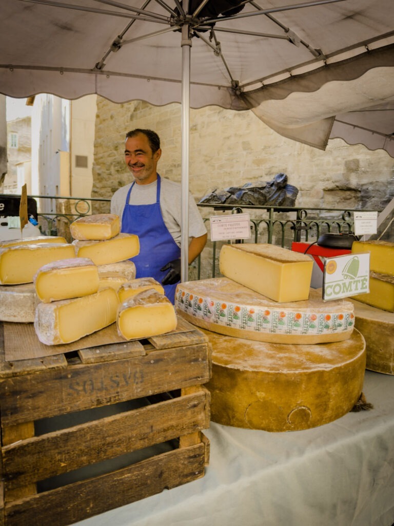 A cheesemonger displaying his local cheeses at an outdoor stall beside the Sorgue River, beneath a white canopy. An example of the type of vendors visitors can discover while browsing in L’Isle-sur-la-Sorgue.