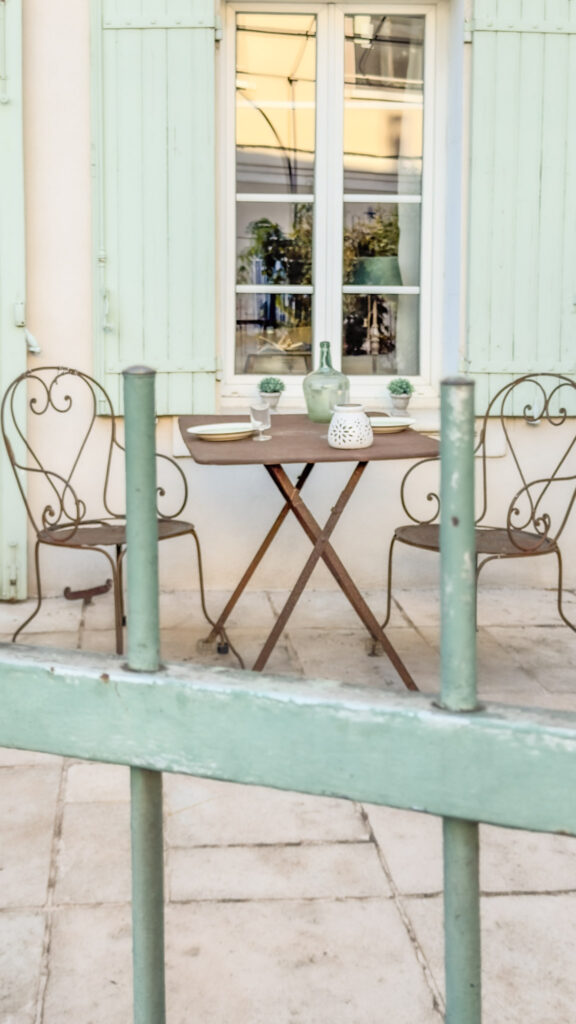Through a green iron gate is a iron table and chairs in front of a window with green shutters at restaurant in L'Isle-sur-la-Sorgue