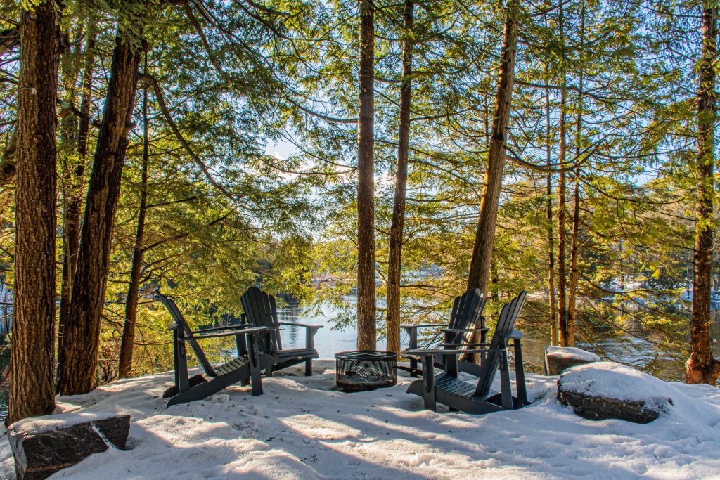 outdoor firepit with gray muskoka chairs with trees and view of lake in winter at Muskoka cottage